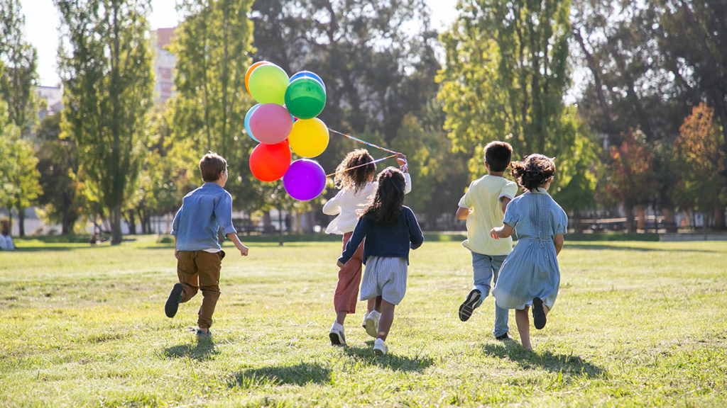 Five children running through a park with one of them holding a bunch of balloons.